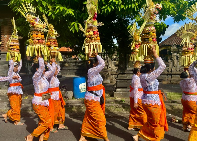 Women in traditional clothing walk along the street in a ceremony. They are balancing offerings of fruit and flowers shaped into a tower.on their heads. They are wearing white lace tops and orange sarons.