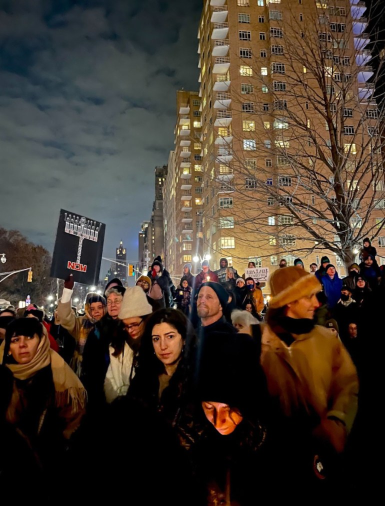 Protesters march against a nighttime sky in New York City, high-rise buildings behind them.