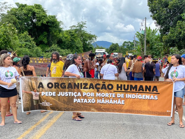 Manifestantes em uma estrada no Brasil seguram uma faixa pedindo justiça no caso de um falecido líder indígena morto a tiros.