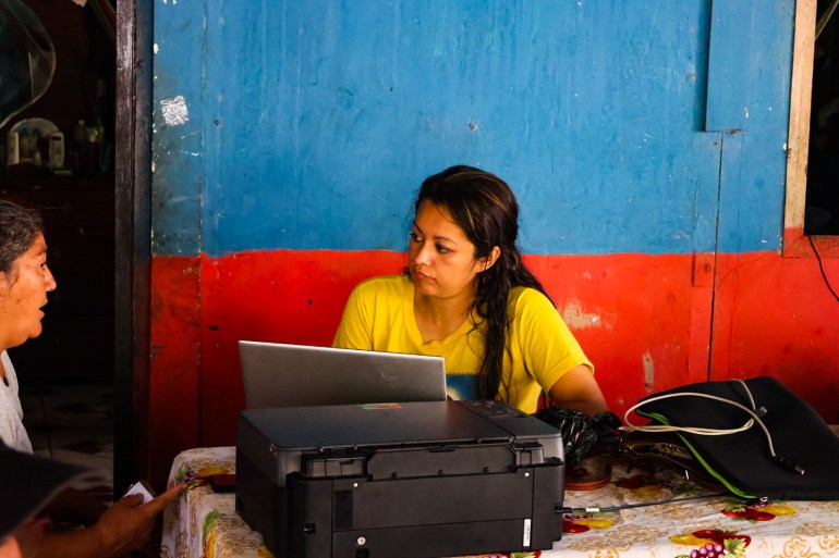 Ingrid Escobar, her long hair in a plait and wearing a yellow T-shirt, sits at table in front of her laptop, as a second woman speaks to her.