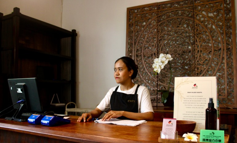 Gerente de spa y terapeuta Murniyati.  Ella está sentada en el mostrador de recepción del spa.  Hay una maceta con orquídeas blancas sobre el escritorio y puertas de madera talladas ornamentadamente detrás.