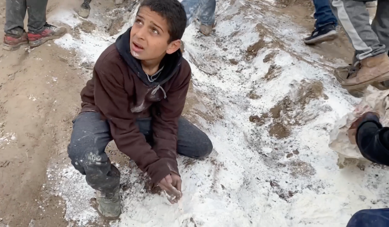 A Palestinian child tries to pick up spilled flour during aid distribution in north Gaza. [Screengrab/Al Jazeera]