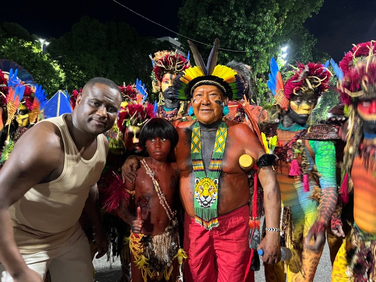 Davi Kopenawa poses for a photo with parade participants at Rio de Janeiro's Sambadrome. He wears a radiating feather headdress, red shorts and a beaded necklace.