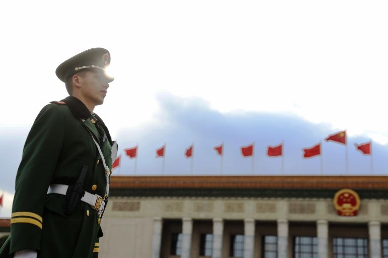 A PLA soldier in profile in front of the Great Hall of the People. Red flags are flying on the roof.