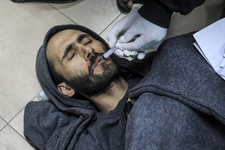 Mohamed Sukkar, eyes closed, lying on the floor of the hospital as someone administers a solution into his mouth using a syringe