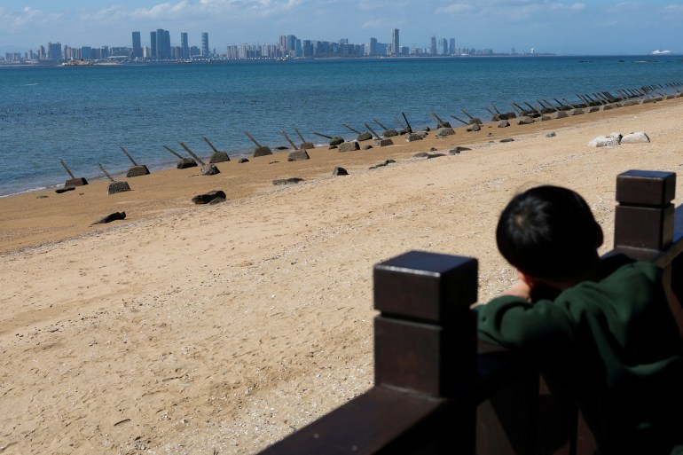 A child looks out at the skyscrapers of Xiamen from the beach on Kinmen. The beach is sandy and the sky is blue.
