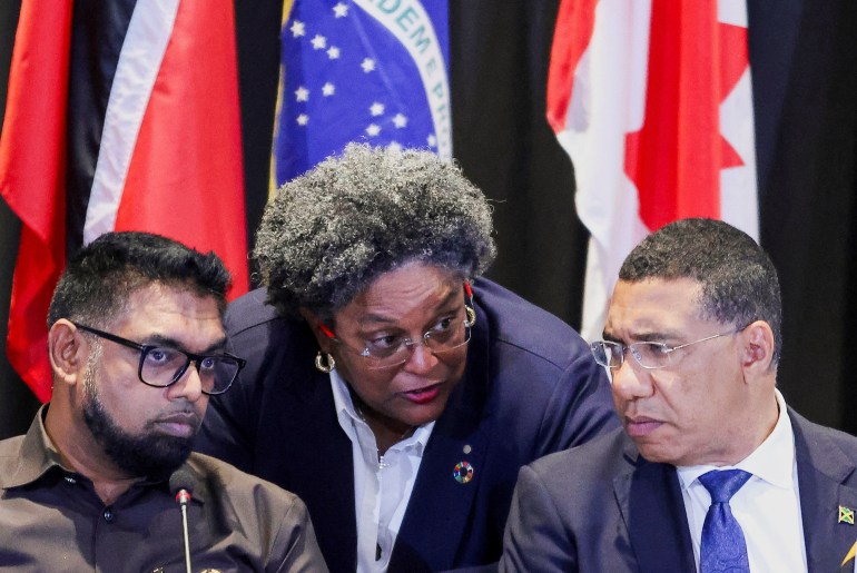 Barbados Prime Minister Mia Mottley speaks with Guyana President Irfaan Ali and Jamaican Prime Minister Andrew Holness. She is in the middle between the two men. There are flags behind them.