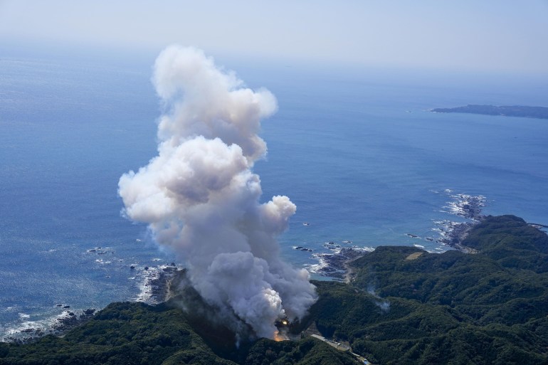 A view of the explosion of the Kairos rocket. There are mountains and sea and clouds of white smoke rising into the sky