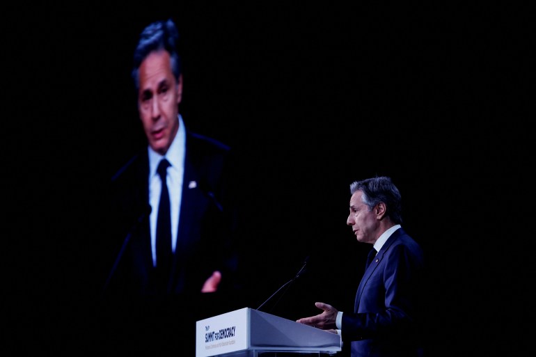a man in a suit speaks in a dark room with a screen showing him on the wall