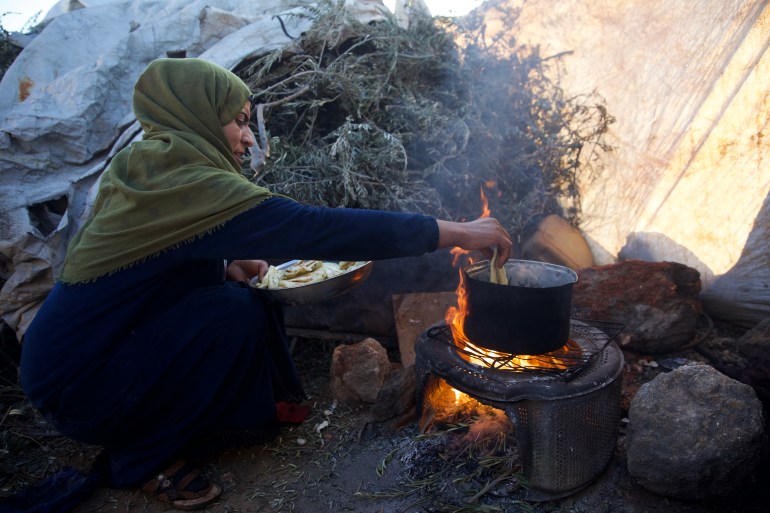 Bayan crouching in front of a fire with a banged-up blackened pot on it, dropping in vegetables to fry