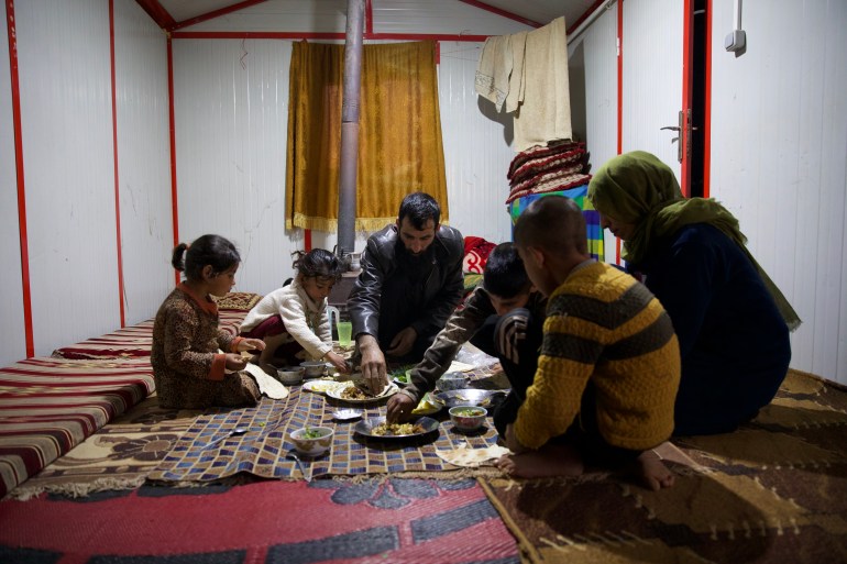 The family gathers around a cloth spread on the floor in a prefabricated shelter