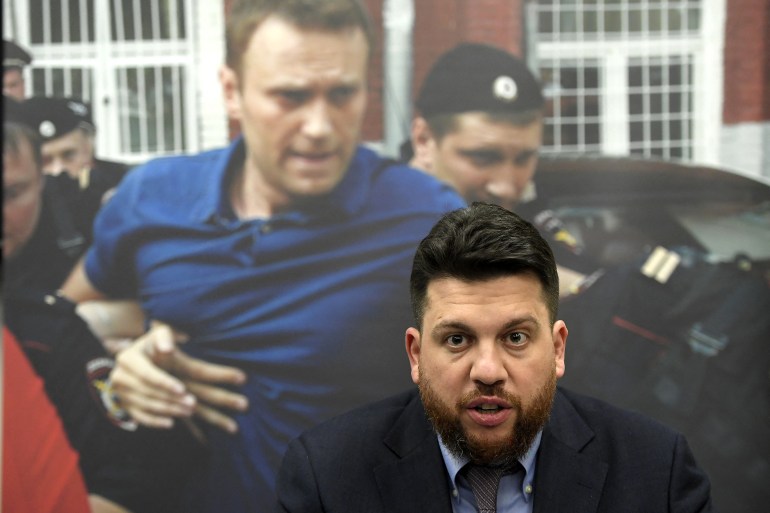 Leonid Volkov speaking at an event at the European Parliament in 2021. He has dark hair and a beard. He is sitting on front of a backdrop showing Alexey Navalny being taken away by police.