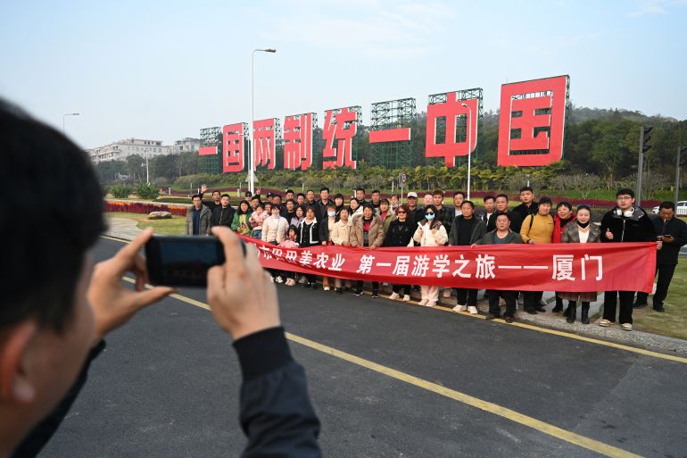 A group of Chinese people take a photo in front of a sign in Xiamen reading 'One country, two systems, unify China'. The words are written in large red Chinese characters.