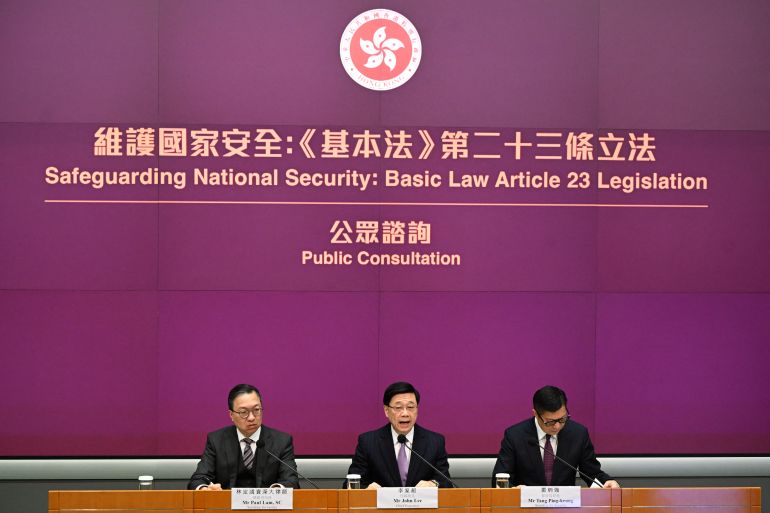 Three top Hong Kong lofficials sitting beneath a purple board reading 'Safeguarding National Security: Basic Law Article 23 Legislation. Chief Executive John Lee is in the middle.
