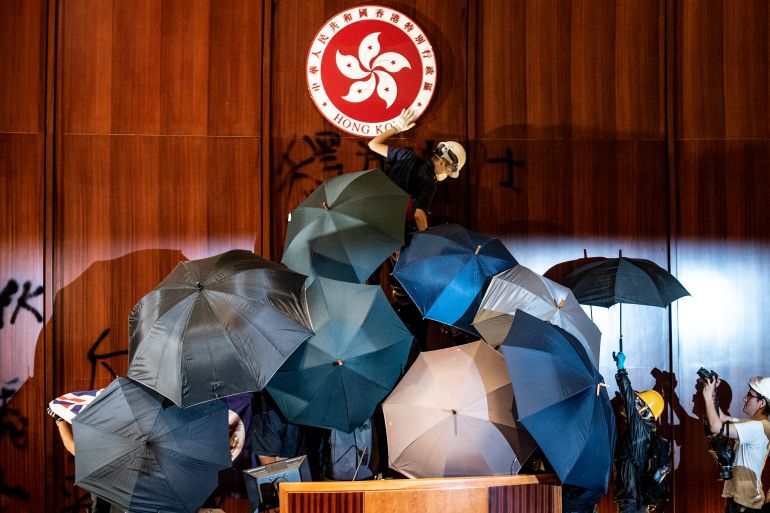 Protesters inside the legislative chamber in 2019. The red and white bauhinia emblem is on the wall above them. The protesters are hidden beneath umbrellas.