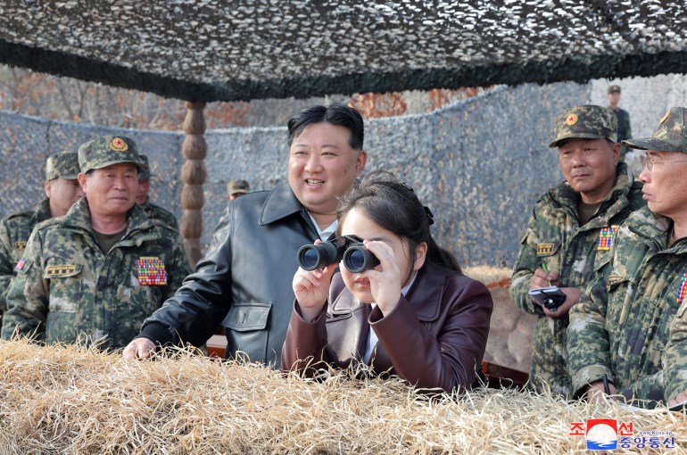 Kim Jong Un with his daughter Kim Ju Ae. They are in a shelter with a number of military officials behind them. Jue Ae is looking through binoculars. Kim is smiling as he stands alongside his daughter.