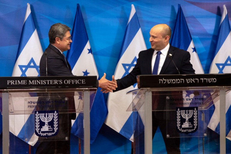 Juan Orlando Hernandez schüttelt dem israelischen Premierminister Naftali Bennett vor einer Reihe israelischer Flaggen die Hand.