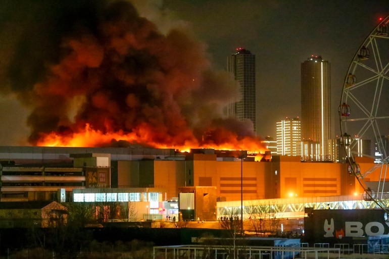 Crocus City Hall engulfed in fire. Orange flames and clouds of black smoke are rising from the roof. The area on the ground is lit up with fire engines. 