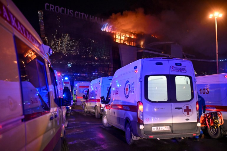Des ambulances font la queue devant l’hôtel de ville de Crocus en feu.  Le lieu est orange en arrière-plan.