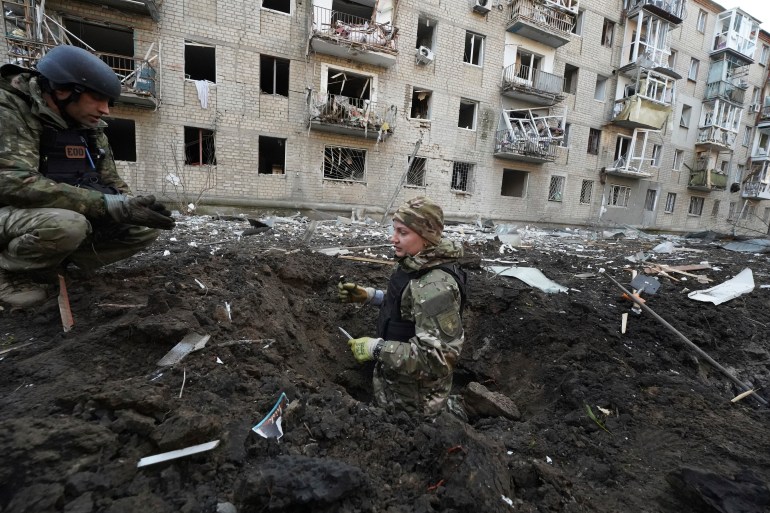 Ukrainian police inspecting a large crater hit by a Russian missile. One is standing in the crater. The other is on the edge of it, squatting down. A damaged apartment building is behind them.