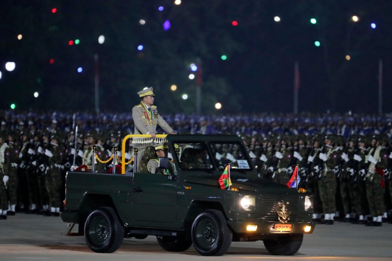 Min Aung Hlaing inspecting troops at the annual Armed Forces Day parade. His is standing on the back of an open-topped jeep-like vehicle. It is night time.