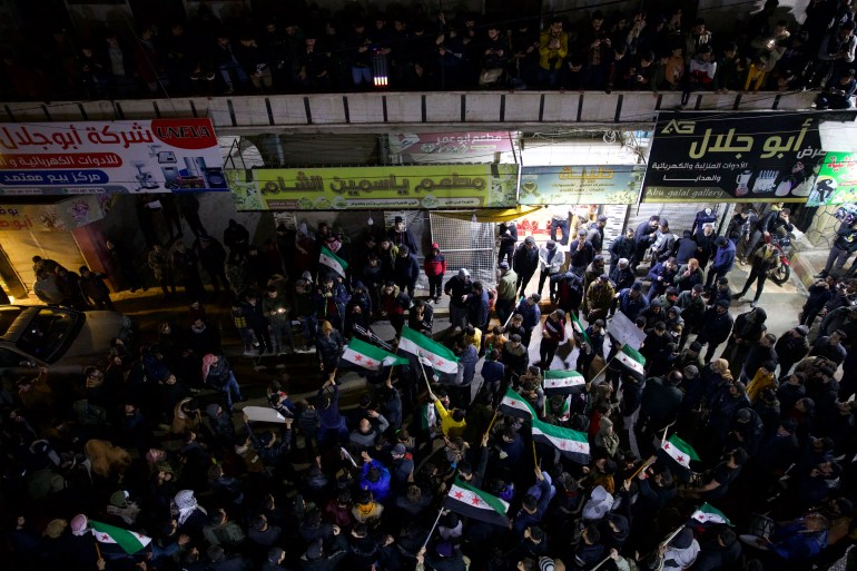 Uma multidão de manifestantes, alguns deles ostentando a bandeira da oposição síria, reúne-se para um protesto.  A foto foi tirada de cima. 