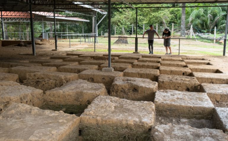 Excavation trenches at the Sungai Batu Archaeological Complex. Large square stones have been unearthed. They are under a canopy, Two people are looking at the site from behind a fence.