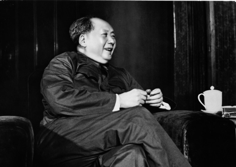 Une photo en noir et blanc de Mao Zedong.  Il est assis et souriant.  Une tasse de thé est posée sur la table à côté de lui.