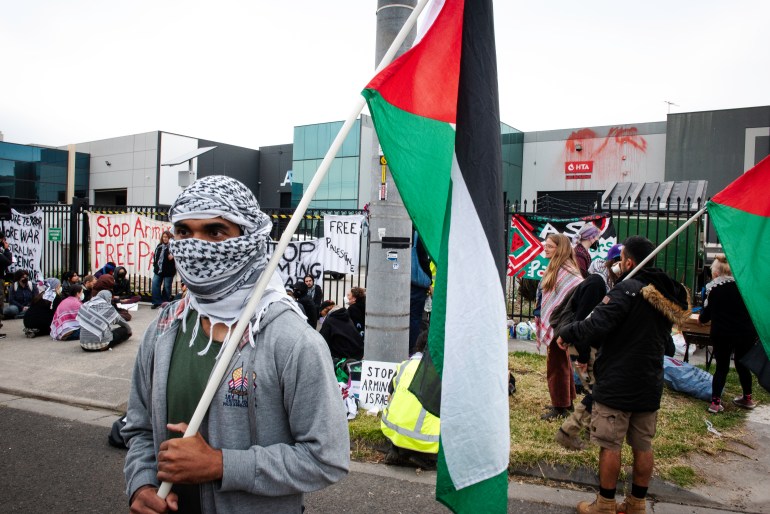 Um manifestante carregando uma bandeira palestina em um piquete em frente a uma empresa de armas australiana.  Eles enrolaram um lenço palestino em volta do rosto para que apenas seus olhos fiquem visíveis. Outros manifestantes estão atrás deles.  Eles têm cartazes.  Alguns estão sentados no chão.