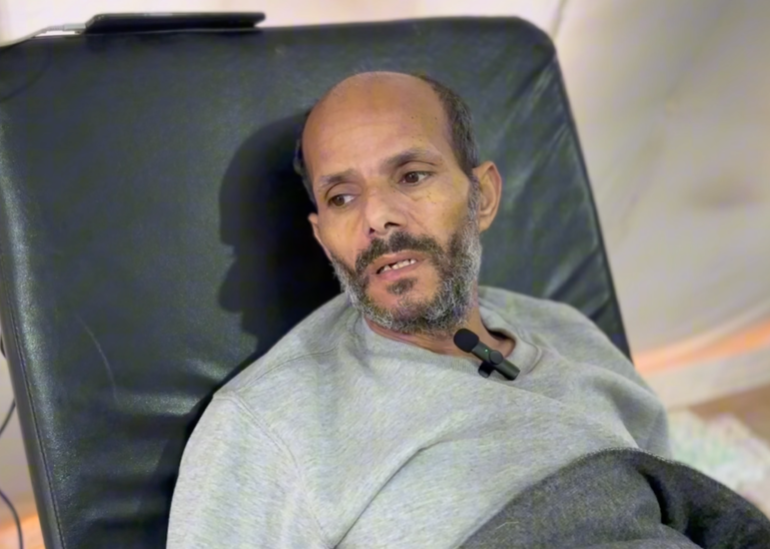 Bald, bearded man lying exhausted on a reclined hospital bench, with a blanket pulled over him