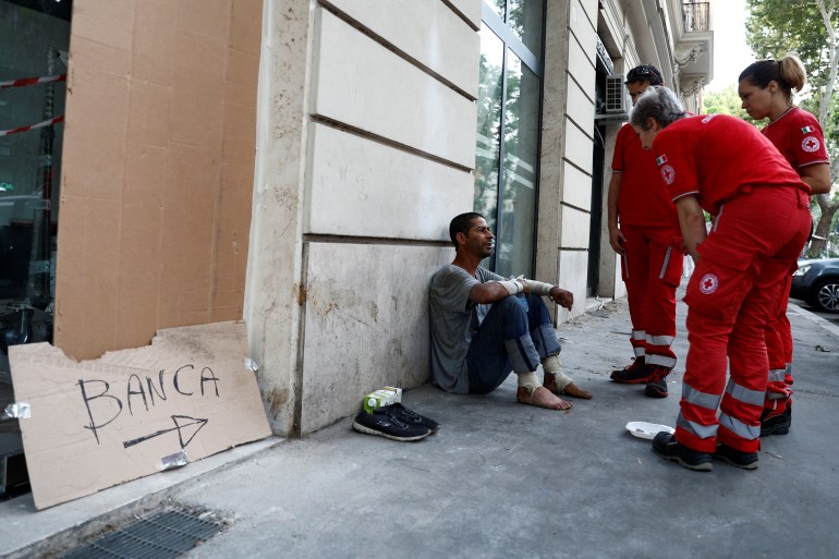 Red Cross workers checking on a homless man during a heatwave in Italy. The man is sitting on the street against a wall. He has taken his shoes off and is barefoot. The three Red Cross workers are in uniforms and speaking to him. 