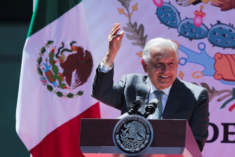 Andres Manuel Lopez Obrador salue derrière un podium, debout à côté d'un drapeau mexicain.