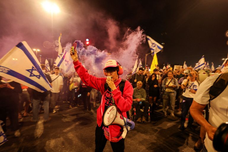 Протестующий в красной куртке дает свисток и держит в правой руке сигнальную ракету. У него громкий грохот. За ними еще больше протестующих. У некоторых есть вспышки. Другие несут израильские флаги.
