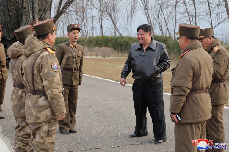 Kim Jong Un com comandantes militares.  Eles estão vestindo uniforme.  Ele está vestido de preto e vestindo uma jaqueta de couro