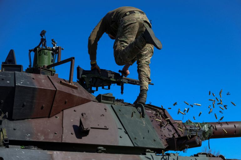 A Marine on top of an armoured vehicle. He is removing empty cartridges during a training exercise. The sky is blue behind him.
