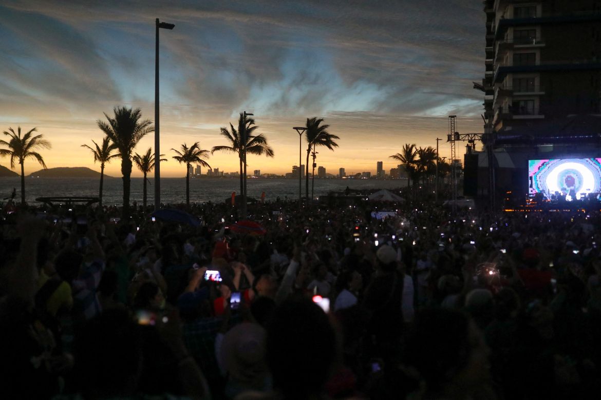 A view of the beaches of Mazatlan, where a crowd darkened by the eclipse hold their phones to the sky. Behind them, a screen and palm trees are visible.