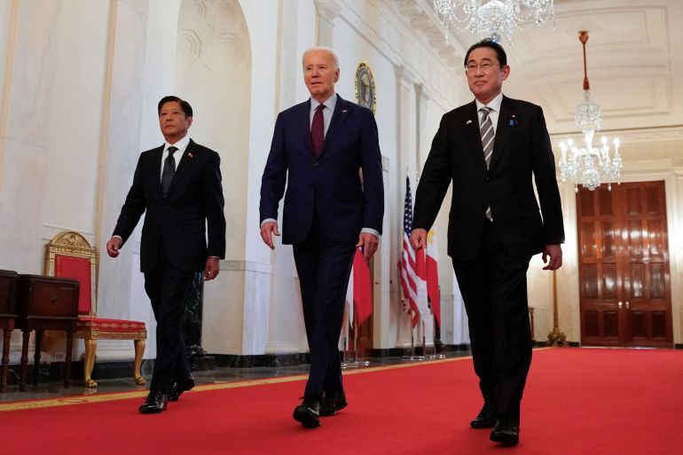 US President Joe Biden escorts Philippines President Ferdinand Marcos Jr and Japanese Prime Minister Fumio Kishida to their trilateral summit at the White House. They're walking along a corridor on a red carpet. They're all wearing dark suits and look relaxed.