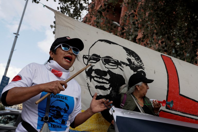 A woman chants while demonstrating in front of a banner with Jorge Glas's face.