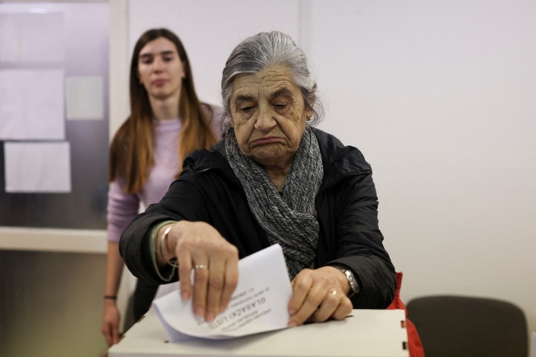 Una donna anziana con i capelli grigi mette la sua scheda elettorale nell’urna.  Sullo sfondo c'è una donna più giovane.