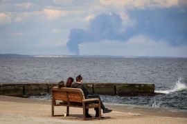 People rest on a beach as smoke rises over the port of Pivdennyi, after a Russian missile strike in Odesa [Nina Liashonok/Reuters]