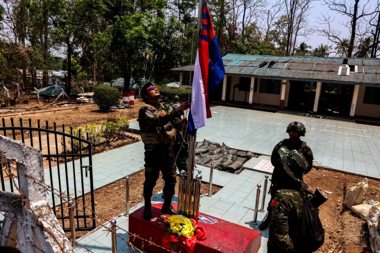 Soldados do KNLA hasteando a bandeira nacional Karen em uma base militar de Mianmar na vila de Thingyan Nyi Naung, nos arredores de Myawaddy.  São quatro lutadores, todos vestindo uniformes de combate.  Eles parecem estar em uma praça de desfile. 
