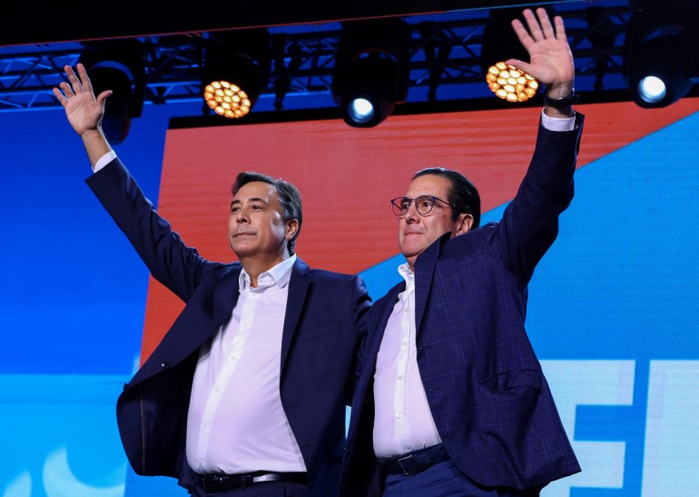 Meliton Arrocha und Martin Torrijos, beide tragen weiße Hemden mit Kragen und dunkle Blazer, umarmen sich und winken dem Publikum bei einer Wahlkampfveranstaltung zu.