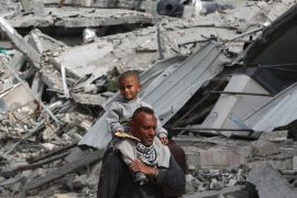 A Palestinian man with his child walk past destroyed buildings in southern Khan Younis after Israel pulled its forces out [AFP]
