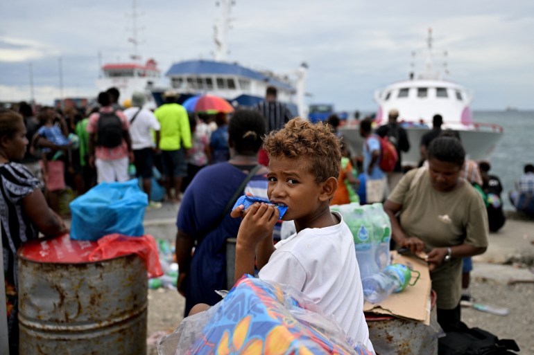 Um menino mastigando um biscoito enquanto as pessoas esperam para embarcar nas balsas para ilhas remotas.