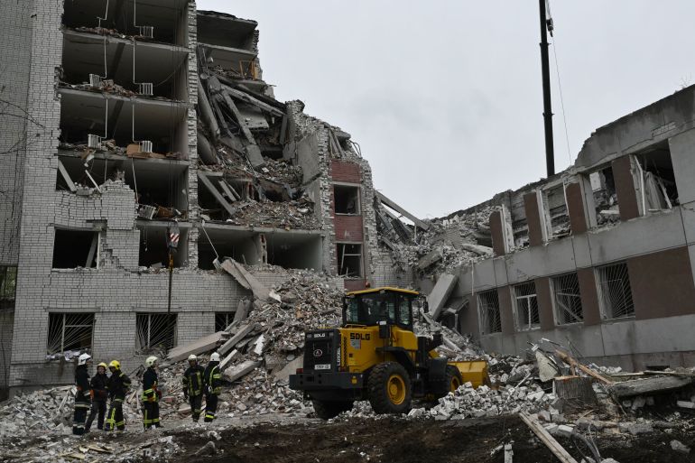 Rescue workers at the site of a collapsed building in Chernihiv. There is an excavator at the front on top of the rubble. The buildings, which are several stories high. are behind.