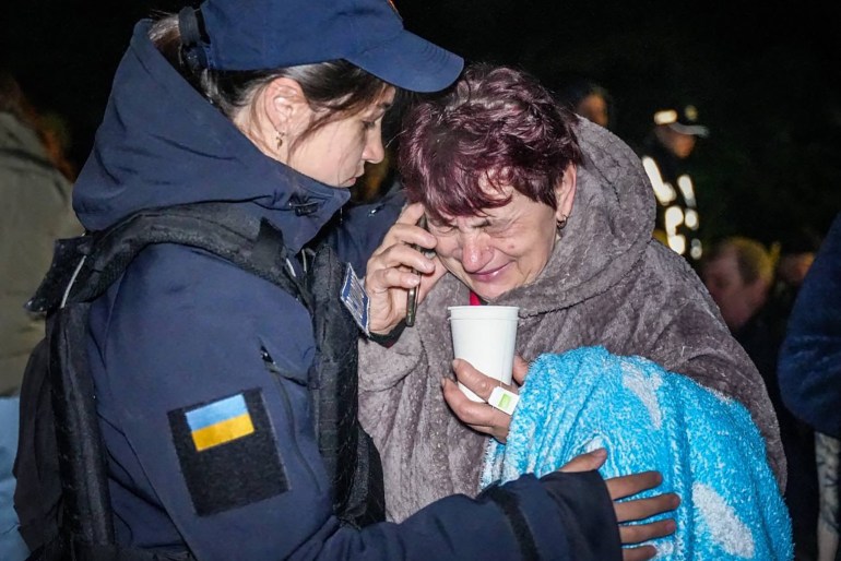 A police officer comforts an older woman in Odesa after a Russian attack destroyed more then a dozen apartments. The older woman is on the phone and clutching a cup of tea. She is sobbing. The officer, a younger woman, is giving her a hug, and looks concerned.