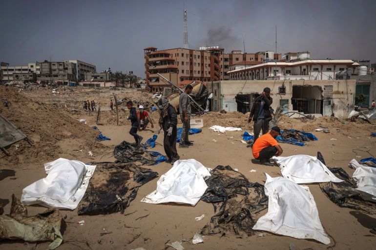 people walk near white sheets lying over bodies next to a destroyed building