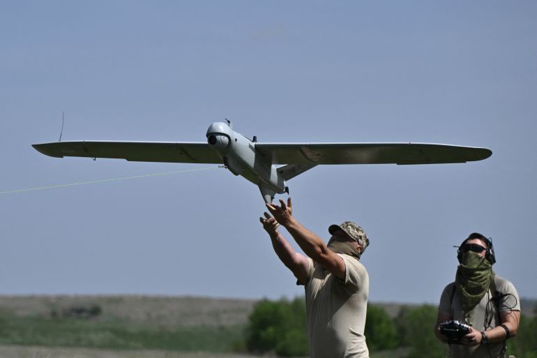 A Ukrainian solider launching a reconnaissance drone. He is holding it up   successful  the air. Another worker  is down  him. The terrain is flat.