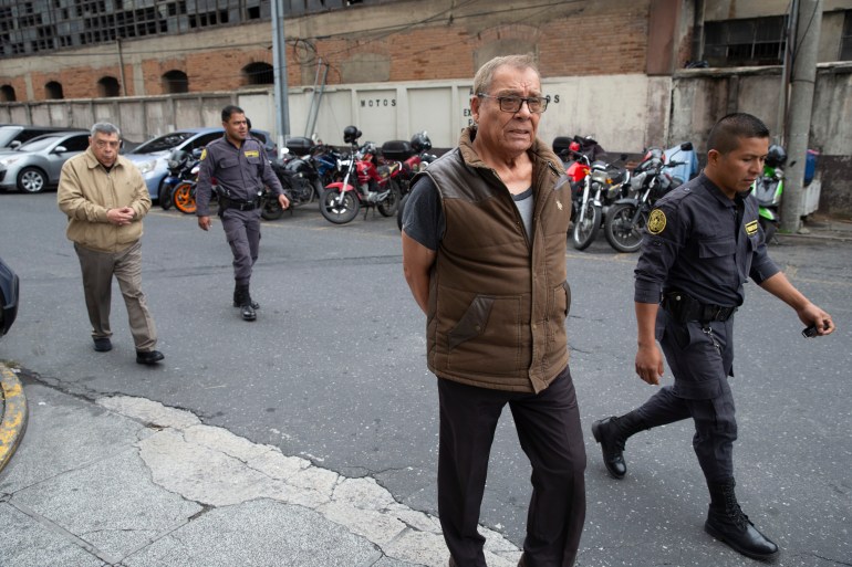 Benedicto Lucas Garcia geht in brauner Weste und Brille durch die Straße.  Hinter ihm sind Strafverfolgungsbehörden zu sehen.