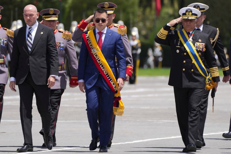 Noboa, wearing a sash and a suit, walks while surrounded by saluting military leaders.